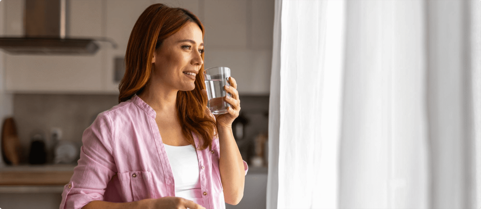 This image shows a person drinking a glass of water while looking out a window.